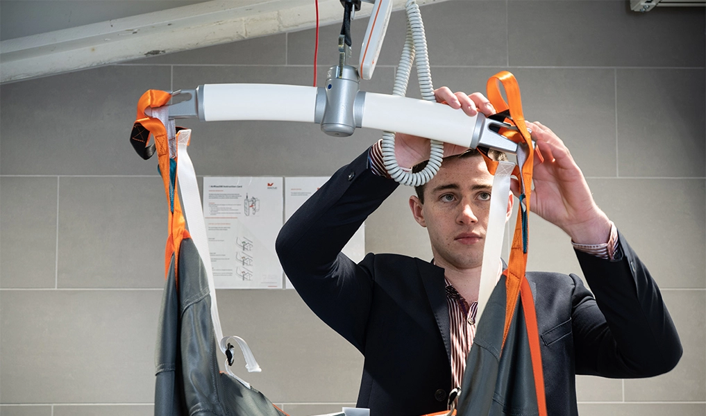 Staff member attaching a hoist sling to a ceiling hoist spreader bar.